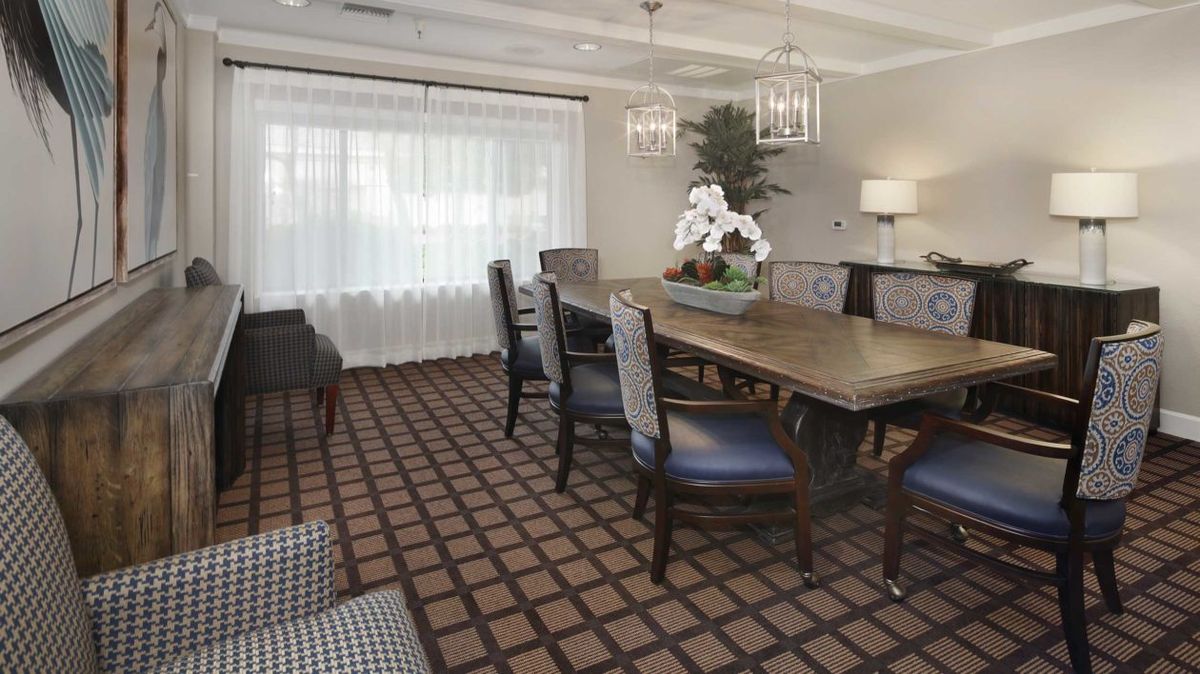 A well-furnished dining or meeting room featuring a large wooden table, upholstered chairs, a patterned carpet, and elegant chandeliers.