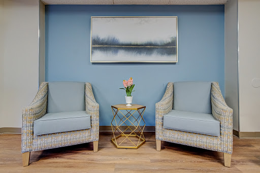 A cozy seating area featuring two light blue chairs, a small gold side table with a flower vase, and a calming piece of artwork on the wall.