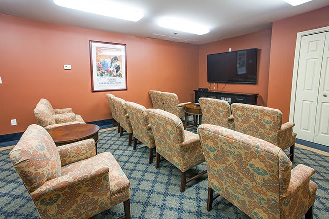 A cozy room with upholstered chairs facing a flat-screen television, adorned with a patterned carpet and a painting on the wall.