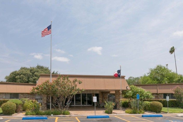 A single-story building with flags and a parking lot in front, surrounded by trees.