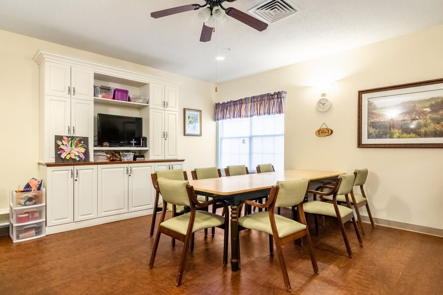 A cozy dining room with a large table and several chairs, featuring a ceiling fan, bookshelf, and window with curtains.