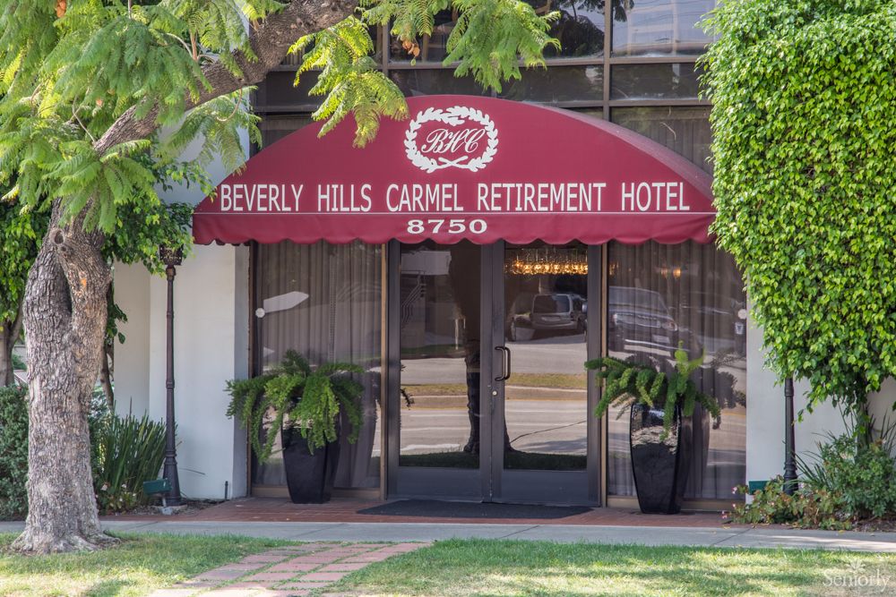 Entrance to the Beverly Hills Carmel Retirement Hotel with a red awning and decorative plants.