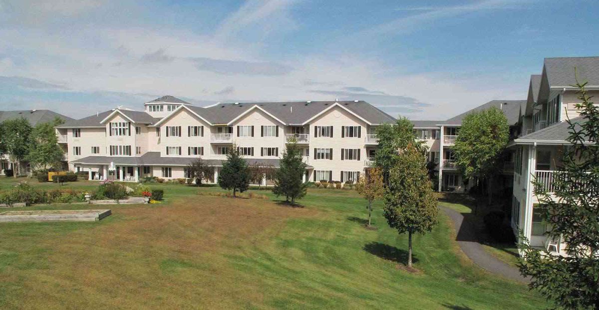 A residential apartment building surrounded by a large lawn and trees under a clear blue sky.
