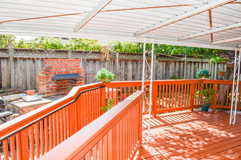 A sunny outdoor patio with a wooden deck, covered by a canopy and featuring a brick grill.