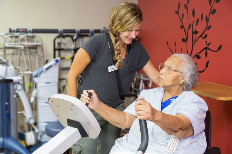 An elderly woman receives assistance from a caregiver during a rehabilitation session involving exercise equipment.