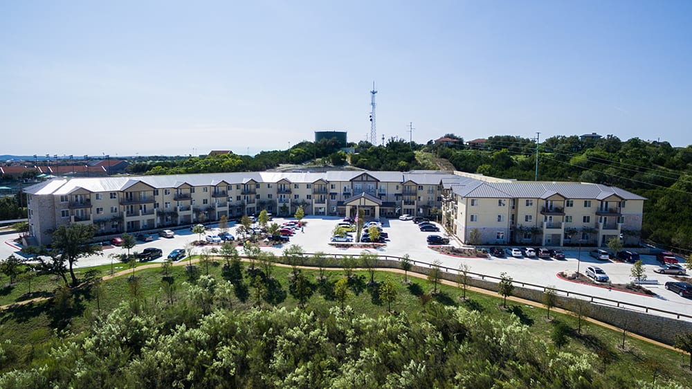 Aerial view of a hotel surrounded by lush greenery and a spacious parking lot.