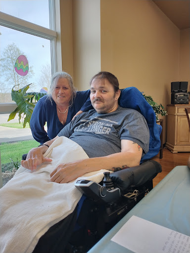 A patient in a wheelchair poses for a photo with a caregiver, both smiling in a bright, airy room.