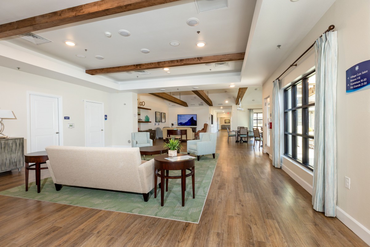 A spacious and well-lit living room featuring modern furniture and wooden flooring, enhanced by natural light and ceiling beams.