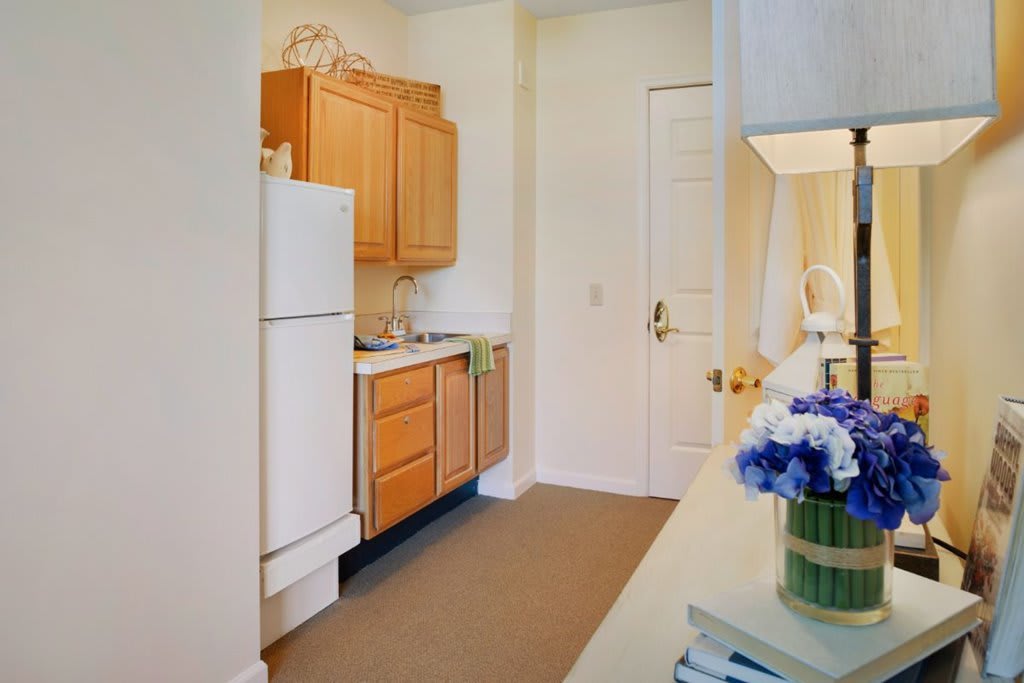 A compact kitchen space with wooden cabinets, a refrigerator, and decorative flowers on the counter.