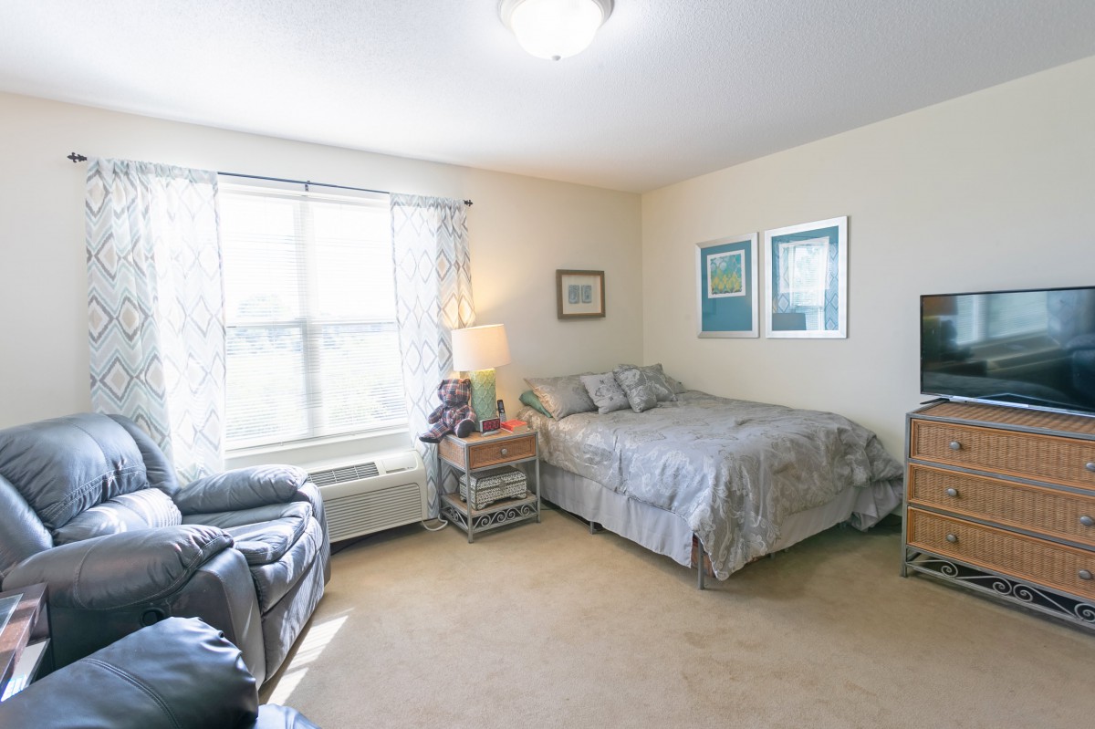 A cozy bedroom with a bed, side table, and armchair, decorated with framed artwork and illuminated by natural light from a large window.