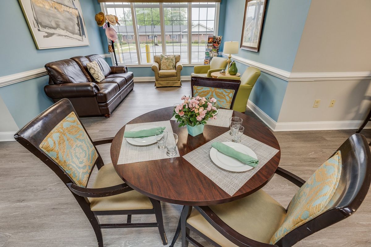 A cozy living room with a round dining table set for two, adorned with flowers, and comfortable seating near large windows.