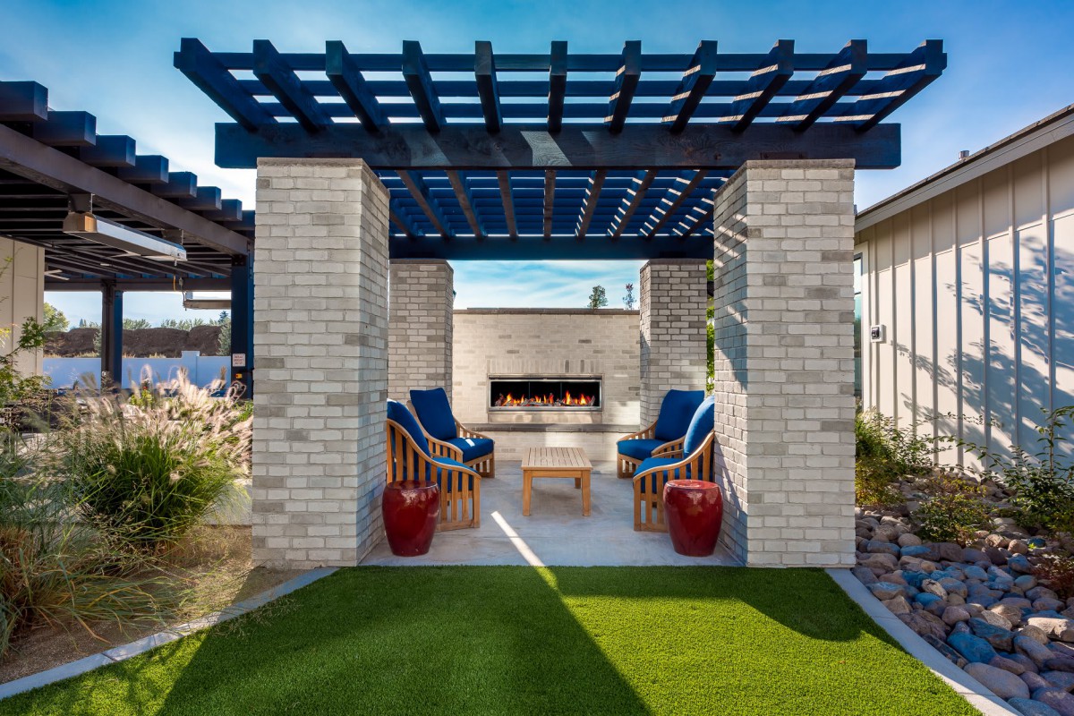 A modern outdoor patio with a pergola and fireplace, featuring cushioned chairs and blue accents.