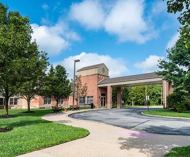 A scenic view of a red-brick building with a circular driveway, surrounded by green trees and under a blue sky with fluffy clouds.