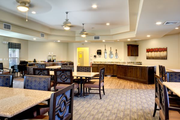 A spacious and well-lit dining area with multiple tables and chairs, featuring ceiling fans and a carpeted floor.