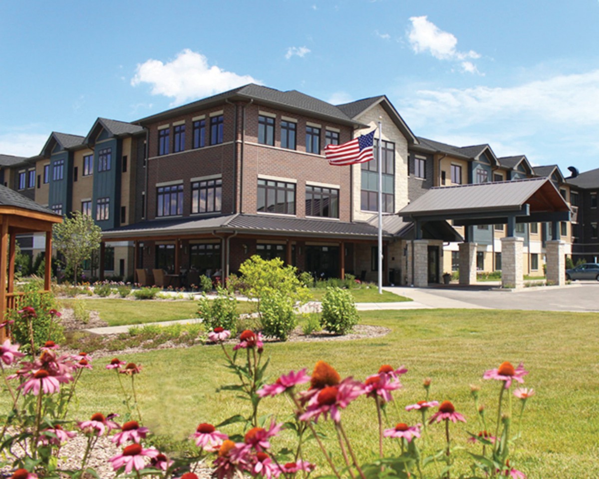 A modern multi-story building with a well-maintained garden featuring colorful flowers and an American flag flying in front.