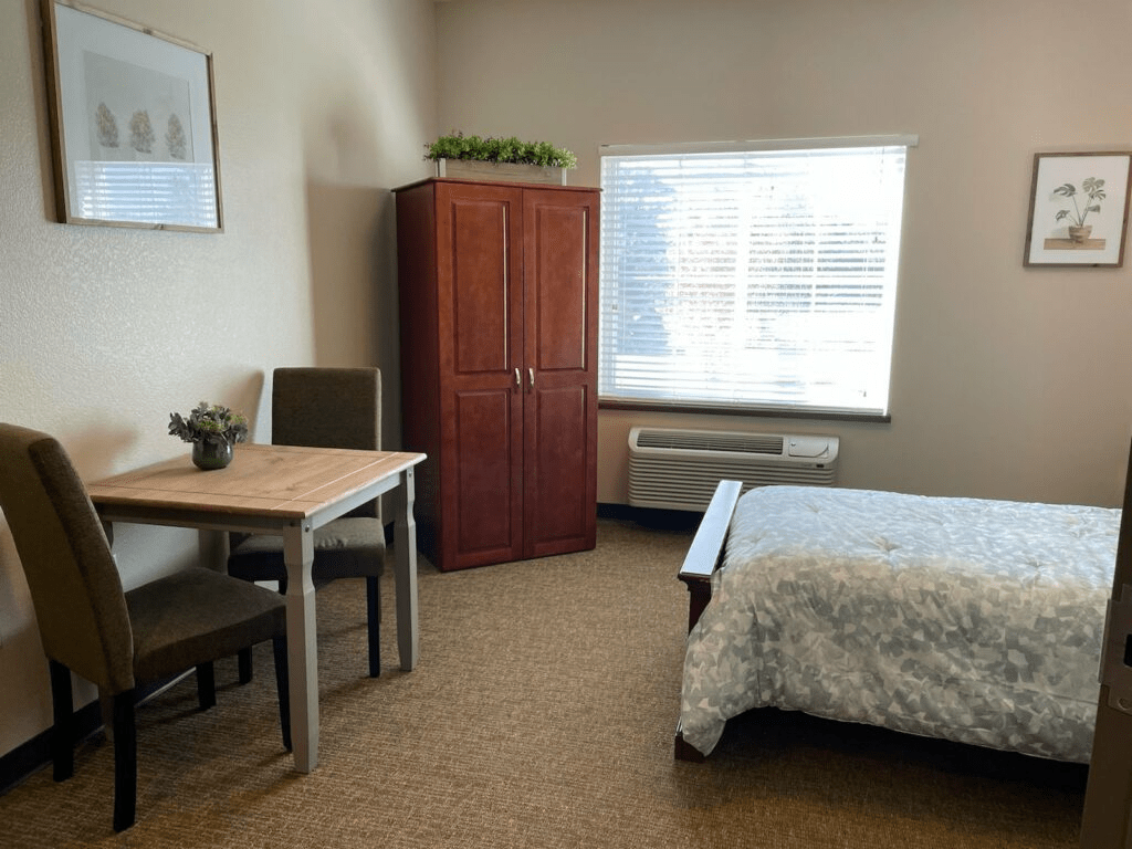 A cozy bedroom with a bed, table, chairs, and a wardrobe next to a bright window.