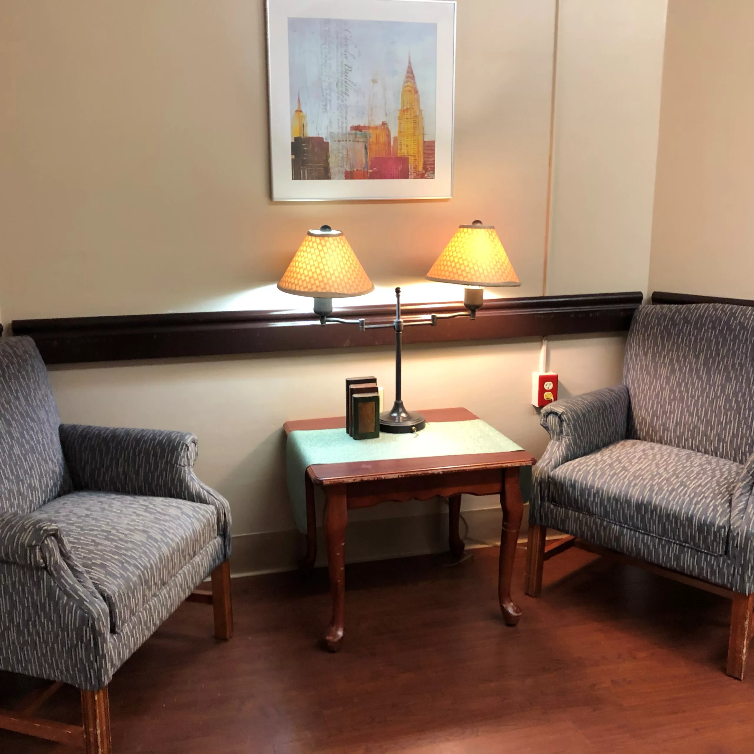 A cozy corner with two upholstered chairs, a wooden table, and a lamp beneath a framed artwork.