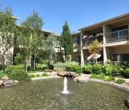 A peaceful apartment complex courtyard with a central fountain surrounded by trees and balconies.