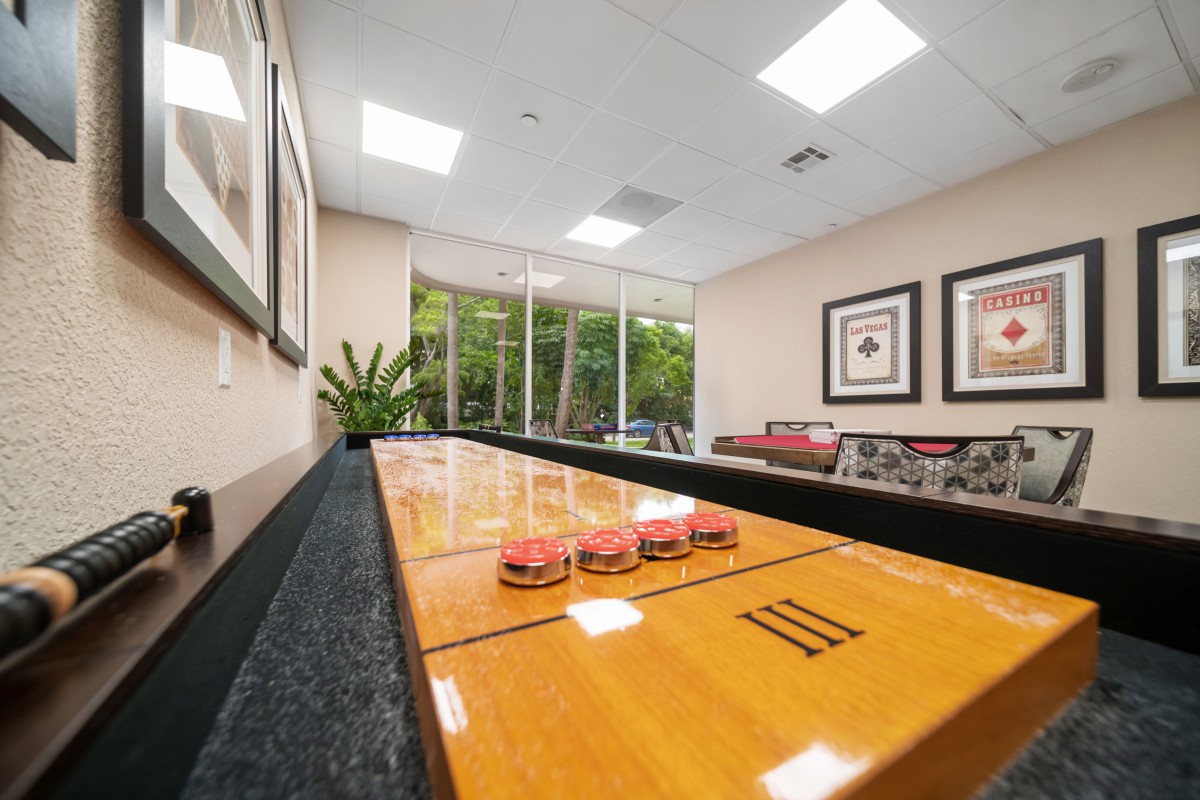 A cozy game room featuring a shuffleboard table, comfortable seating, and large windows overlooking greenery.