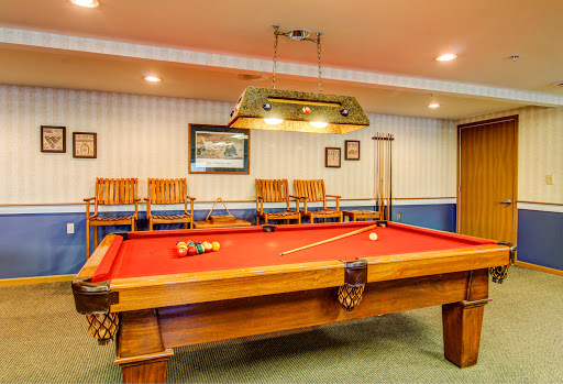 A well-lit game room featuring a classic pool table with red felt, surrounded by wooden chairs.