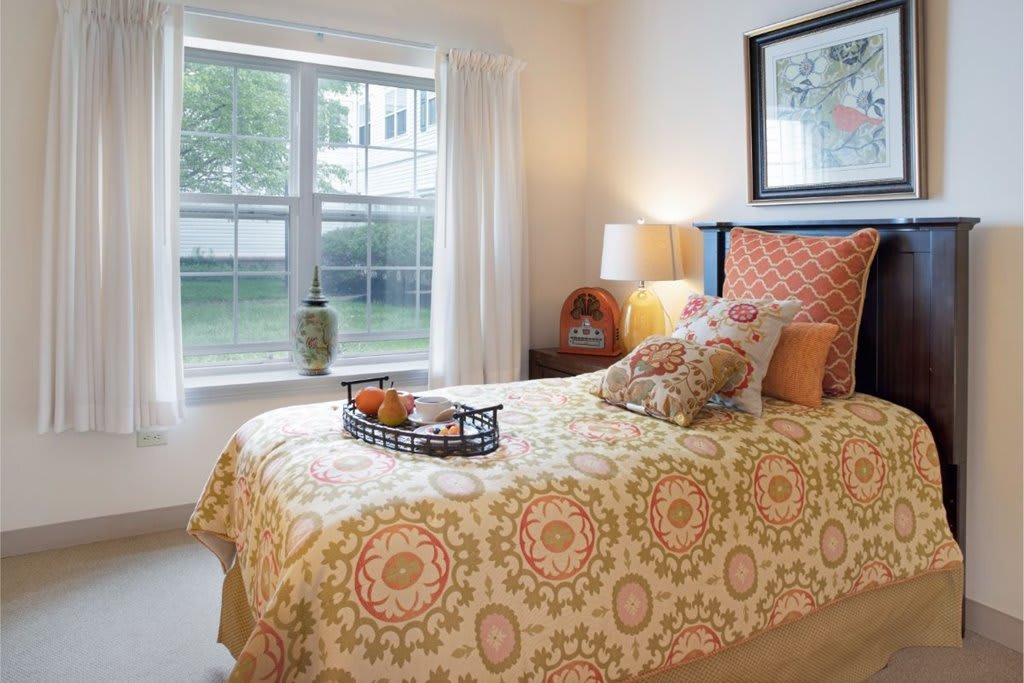A cozy bedroom with a neatly made bed, a bedside lamp, and a window letting in natural light.