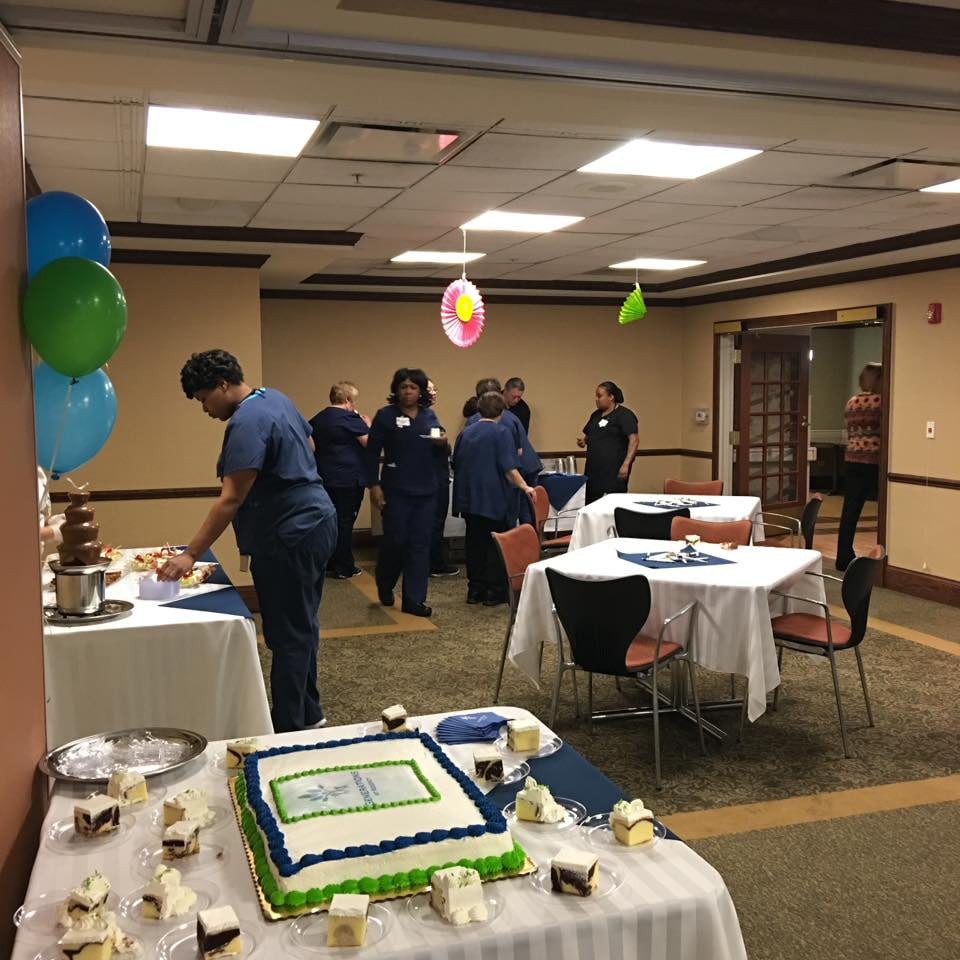 A group of people gather in an indoor setting for a celebration, with balloons and a decorated cake on display.