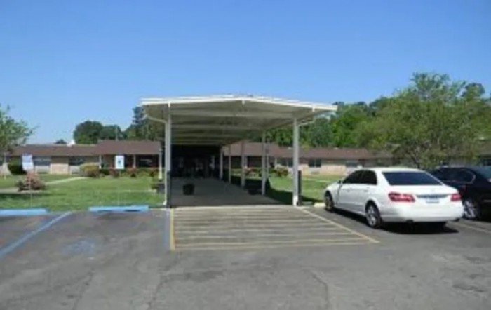 A car is parked under a carport in front of a building with surrounding greenery.