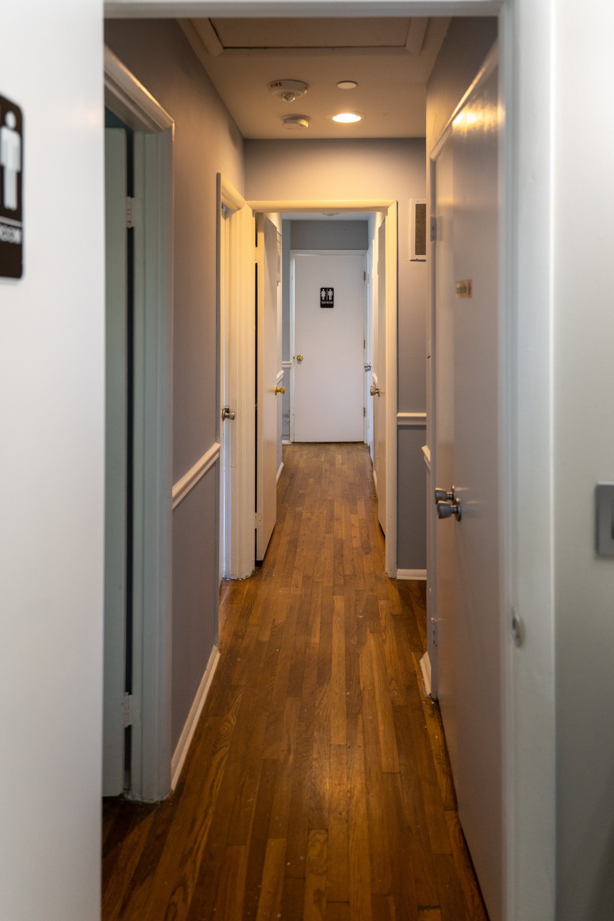 A narrow, well-lit indoor hallway with wooden flooring and multiple doors leading off the corridor.