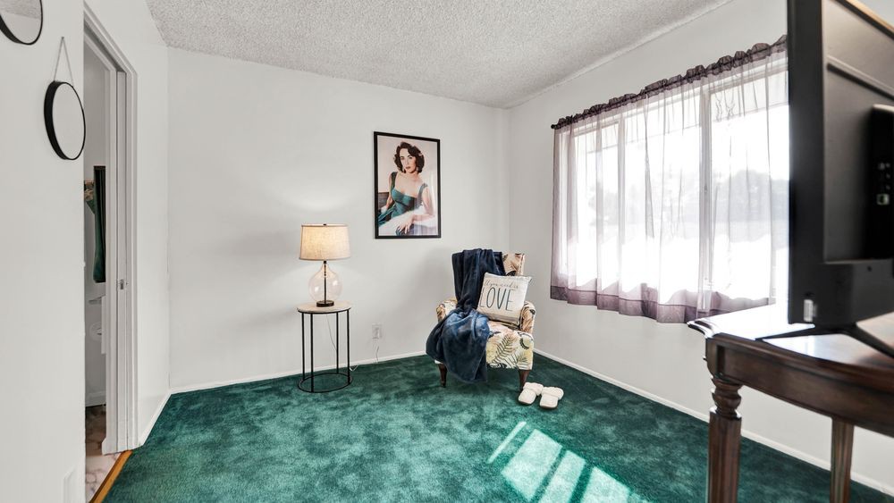 A cozy living room corner featuring an armchair, a small side table with a lamp, green carpeting, and a large window with curtains.