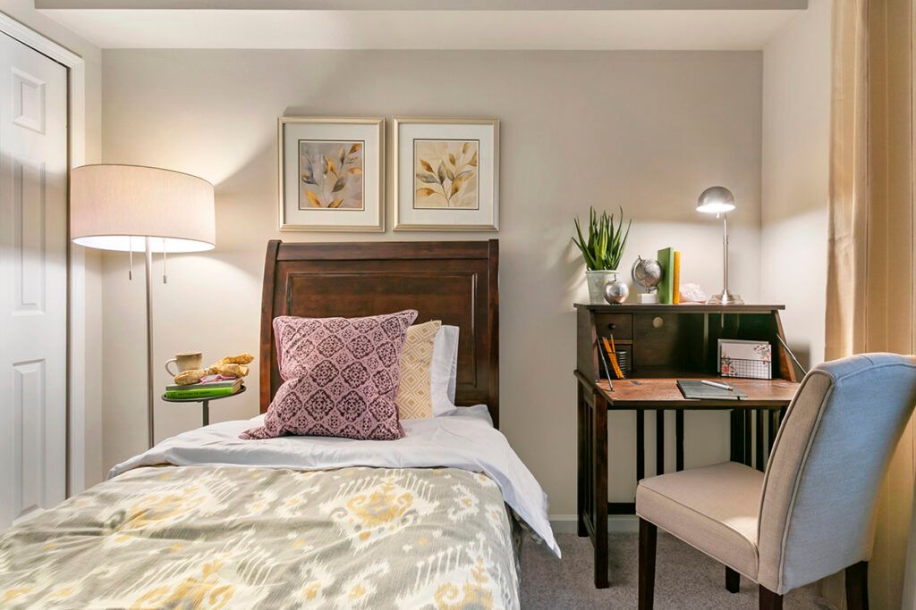 A cozy bedroom with a single bed, decorative pillows, a desk with a lamp, and framed artwork on the wall.