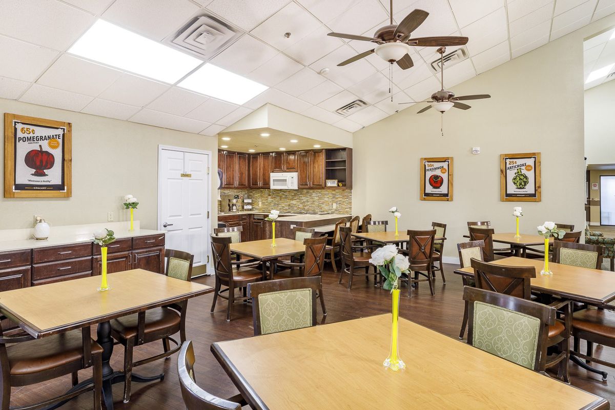 A dining room with several tables and chairs, featuring a kitchen area in the background and ceiling fans overhead.