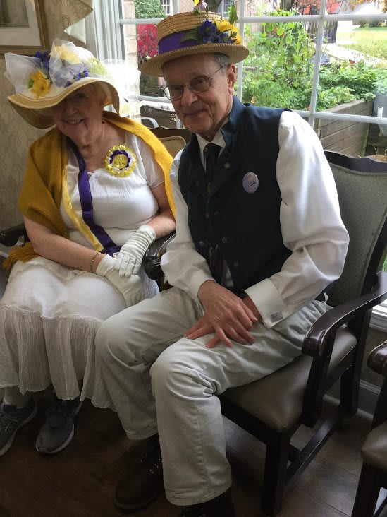 An elderly couple dressed in formal attire sits together outdoors.