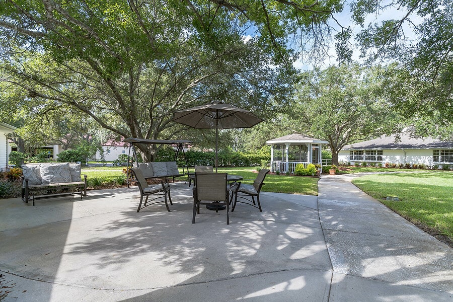 A serene outdoor patio area with tables and chairs set under large trees, featuring a nearby gazebo.