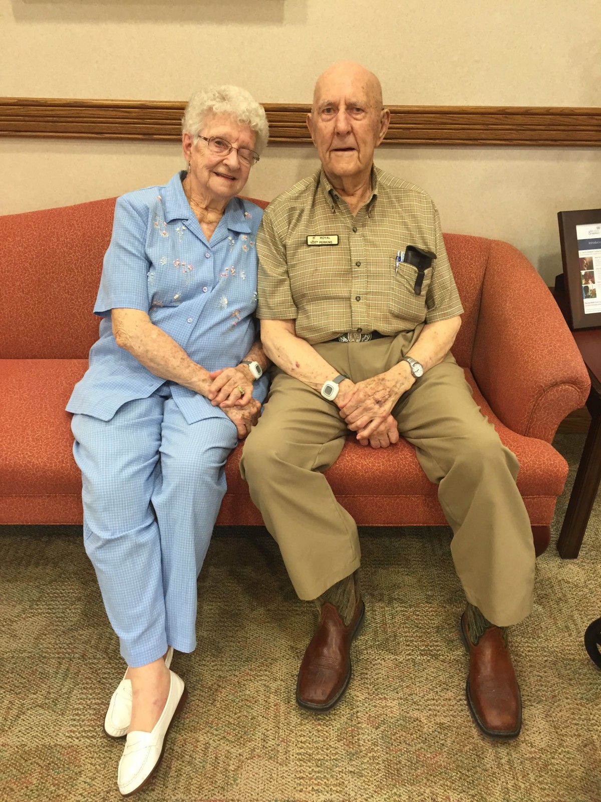 An elderly couple sitting together on a couch, smiling and holding hands.