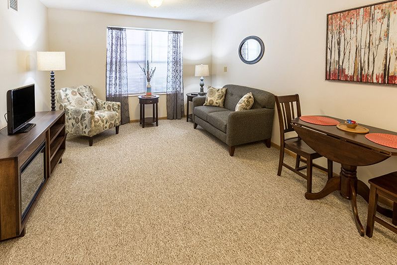 A cozy living room with a sofa, armchair, and a table, decorated with a carpet and lamp.
