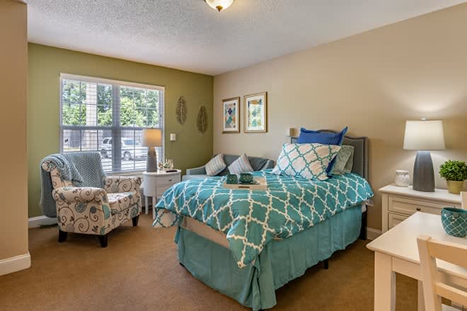 A cozy bedroom with a neatly made bed, decorated with blue and white bedding, a comfortable chair, and a large window letting in natural light.