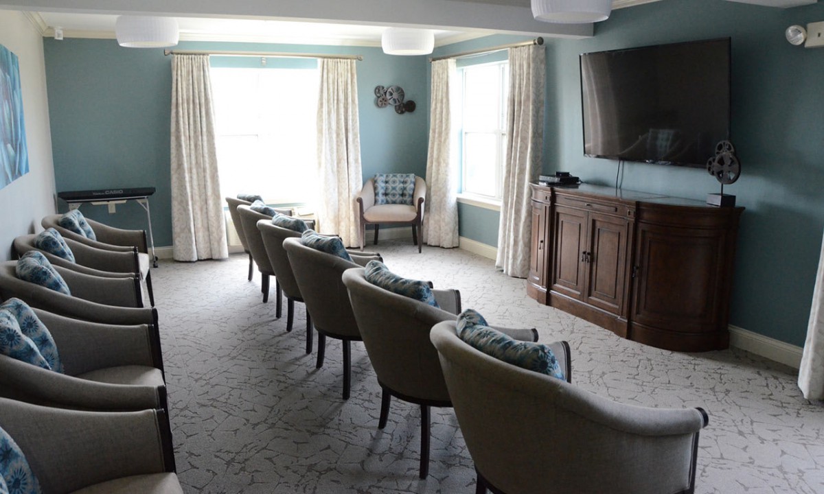 A cozy living room with rows of chairs facing a television, featuring large windows and elegant curtains.