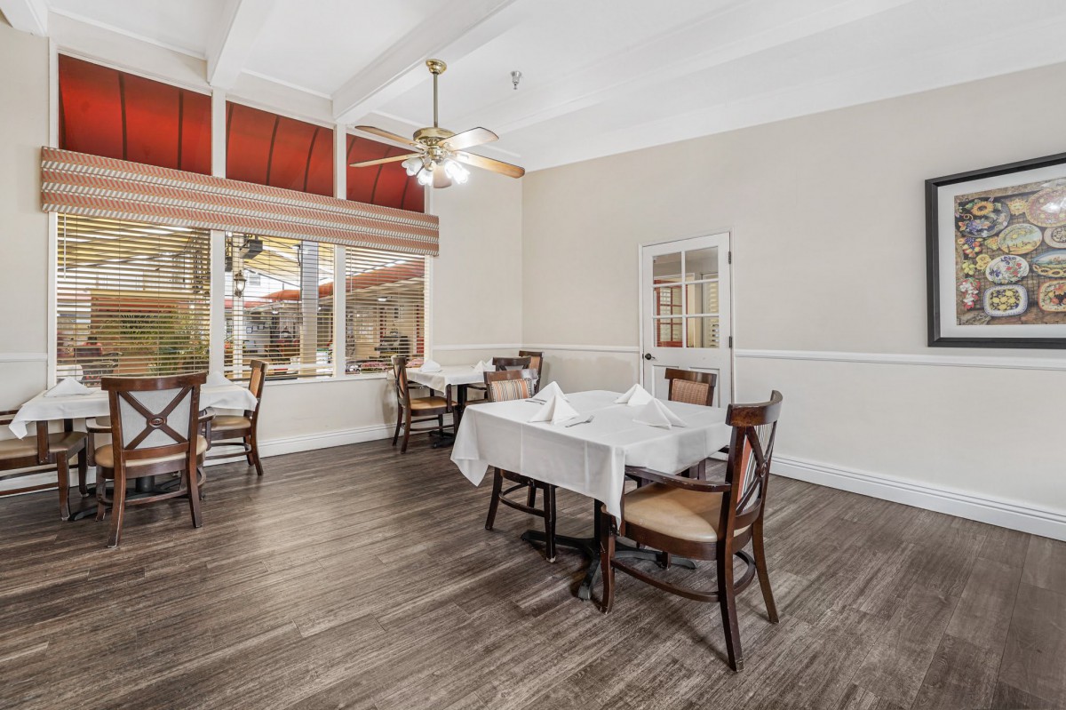 A well-lit dining room featuring wooden floors, tables with white tablecloths, and a ceiling fan.