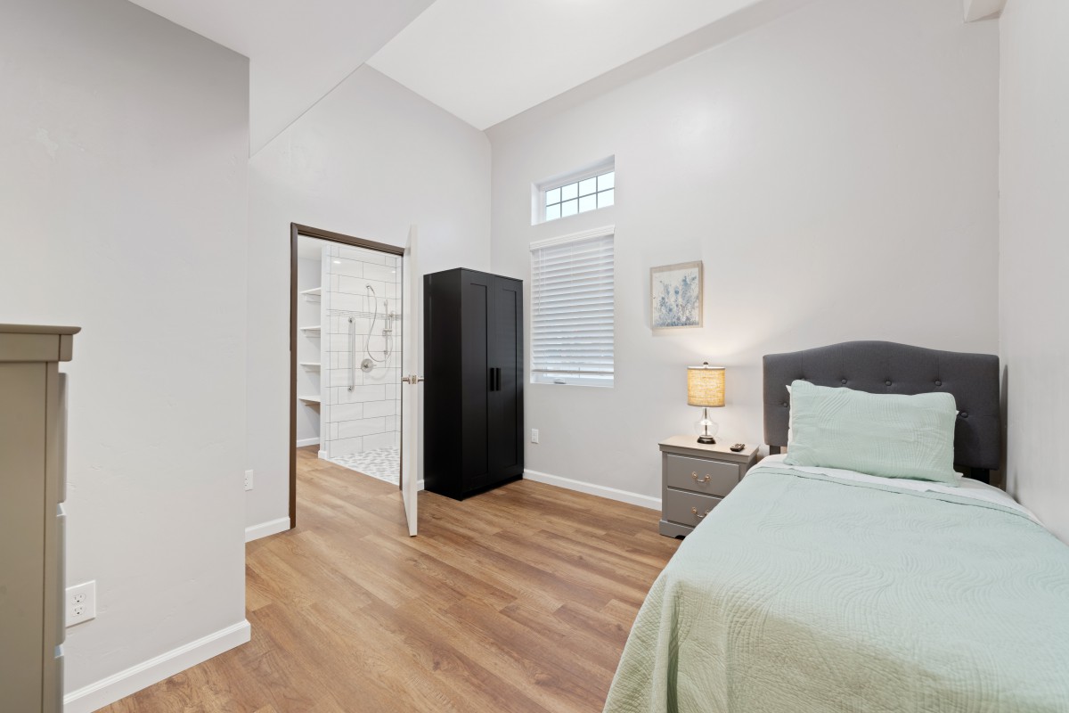 A modern, minimalist bedroom with wood flooring and simple furnishings, including a bed, nightstand, lamp, and wardrobe.