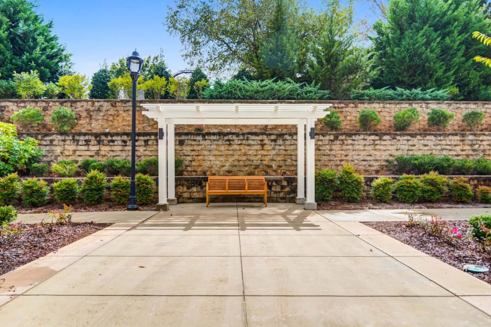 A wooden bench under a white pergola against a brick wall, surrounded by greenery and lit by a nearby lamp post.