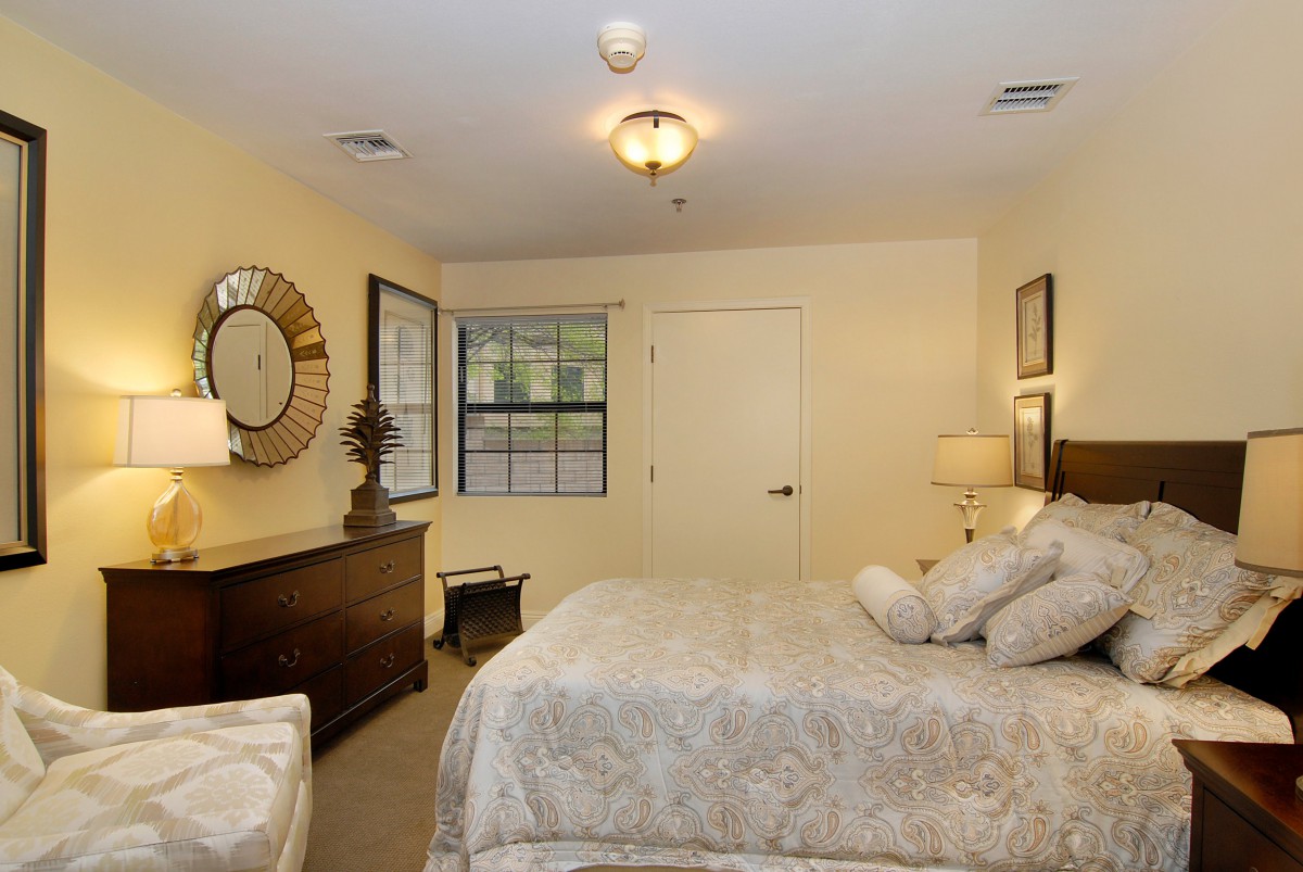 A cozy bedroom with elegant decor, featuring a large bed, mirror, window, and warm lighting.