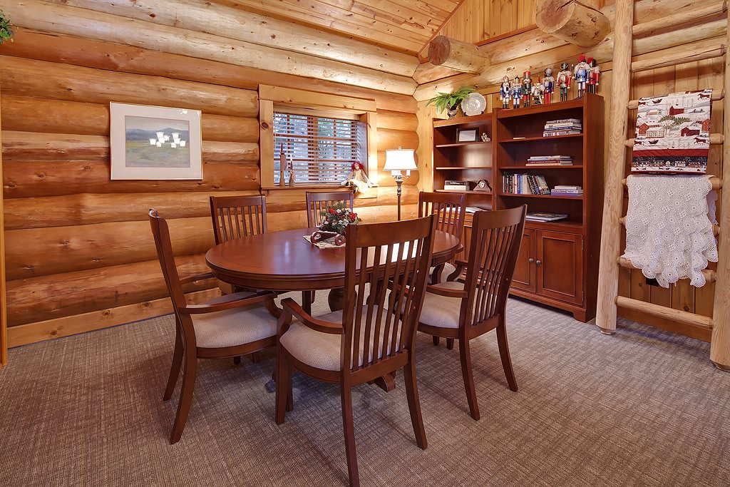 A cozy log cabin dining room with a wooden table and chairs, surrounded by warm wood-paneled walls and a well-stocked bookshelf.