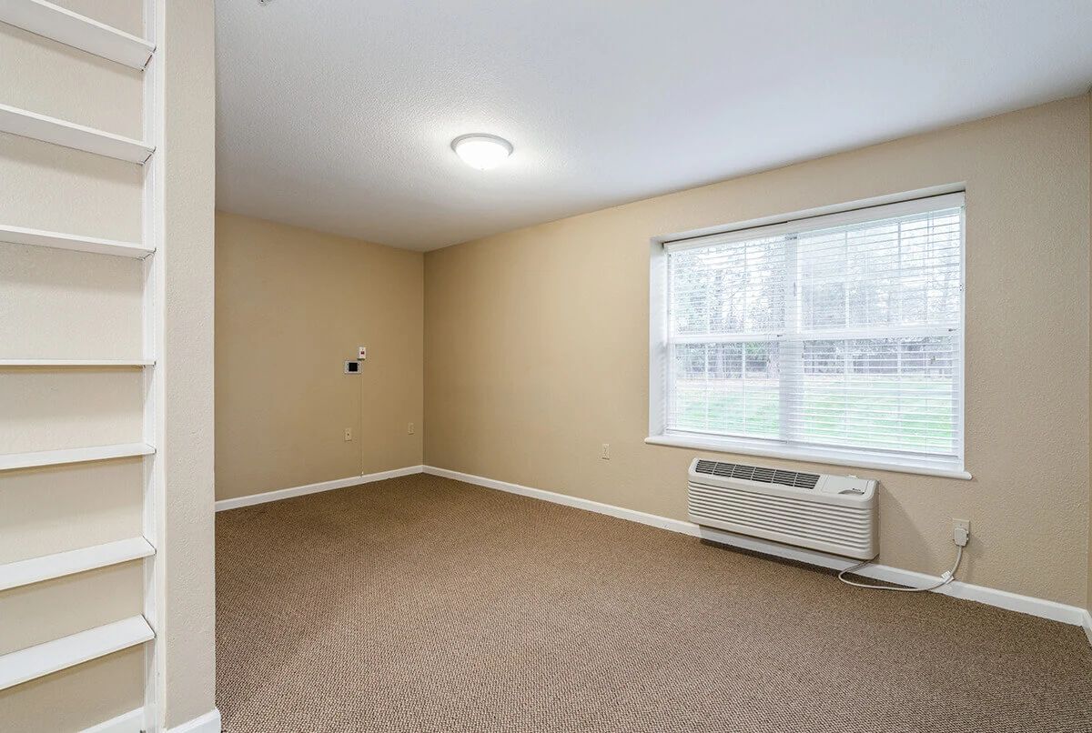 An empty room with beige walls, carpet flooring, a window, and an air conditioner.