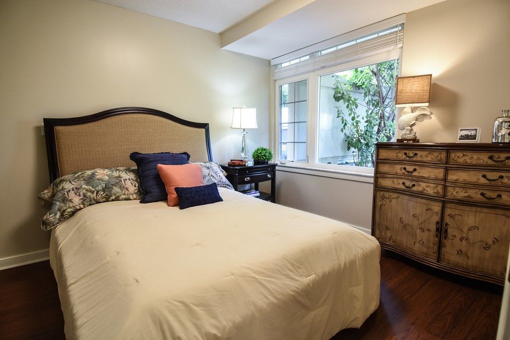 A cozy bedroom with a neatly made bed and decorative pillows, featuring a bedside lamp and a window with greenery outside.