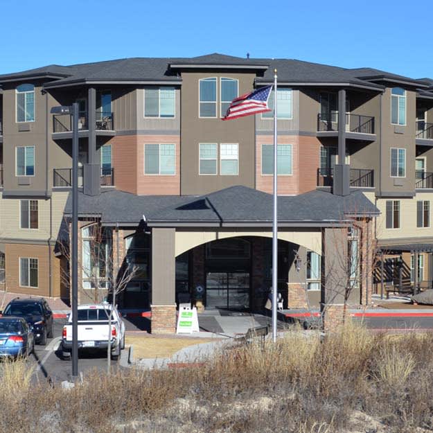 A modern apartment building with a flagpole displaying the American flag, surrounded by parked cars.