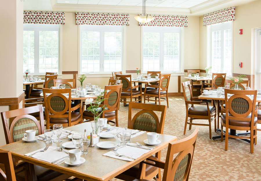 A well-lit dining room with neatly set tables and wooden chairs, featuring large windows that allow natural light to fill the space.